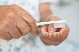 Hands brushing a set of dentures with a basic white toothbrush